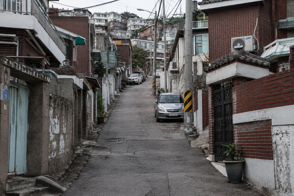 A steep residential alley in Seoul lined with older brick and concrete homes, utility wires, parked cars, and small potted plants